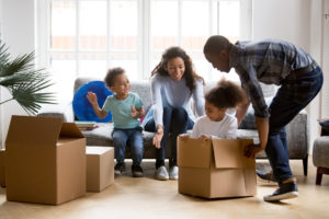 Happy playful large African American family moving in new apartment, little preschooler daughter sitting in cardboard boxes, father rolling her to mother, playing together, purchase property concept