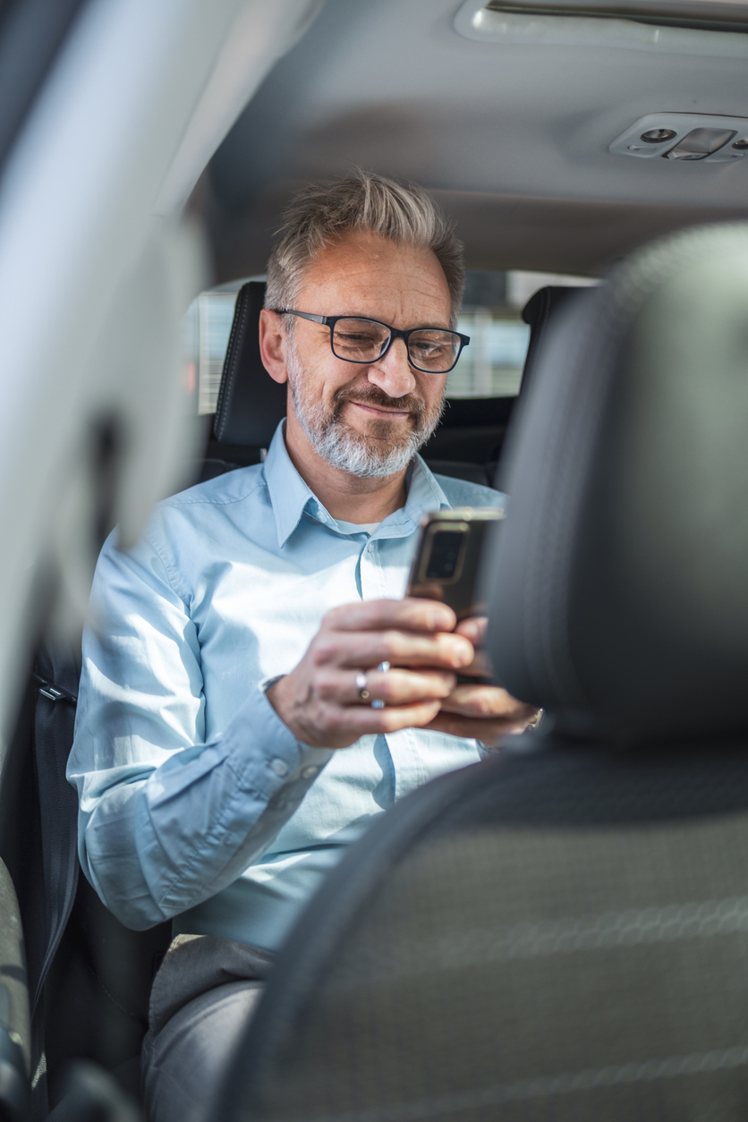 Mature Businessman Commuting In A Taxi While Working Remotely. He Is Focused On His Smartphone, Wearing Glasses, And Dressed In A Light Blue Shirt. Concept Of Business Travel And Mobile Working.