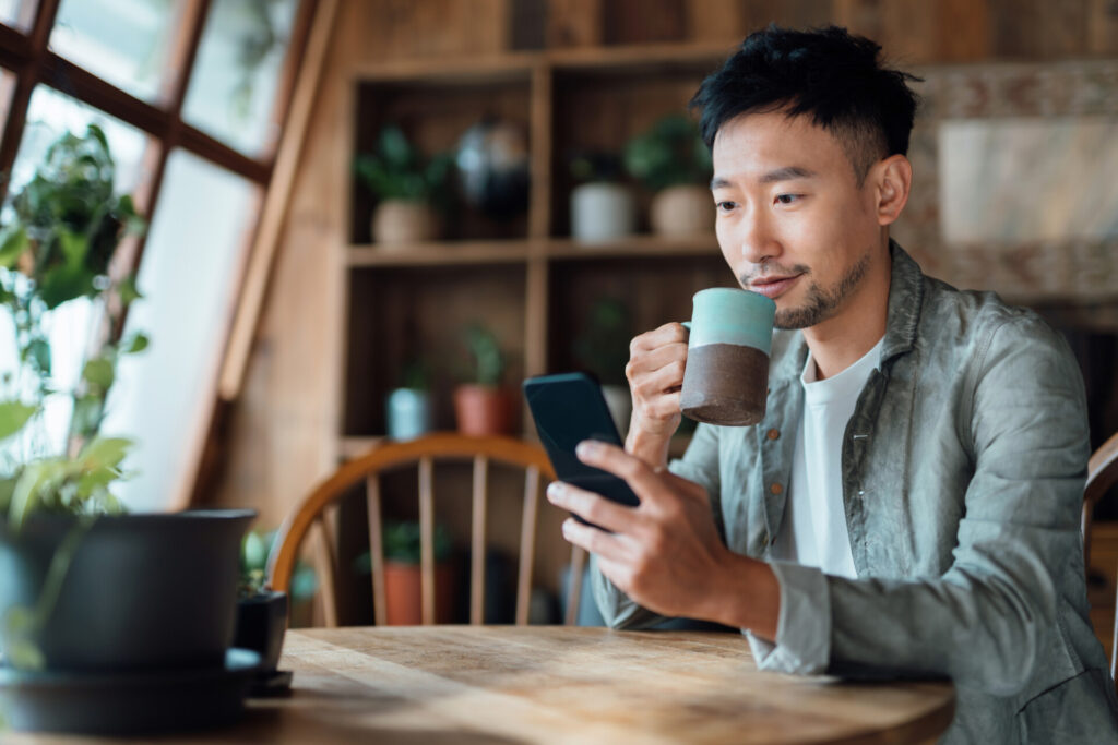 Young Asian Man Managing Online Banking With Mobile App On Smartphone, Taking Care Of His Money And Finances While Relaxing At Home. Banking With Technology